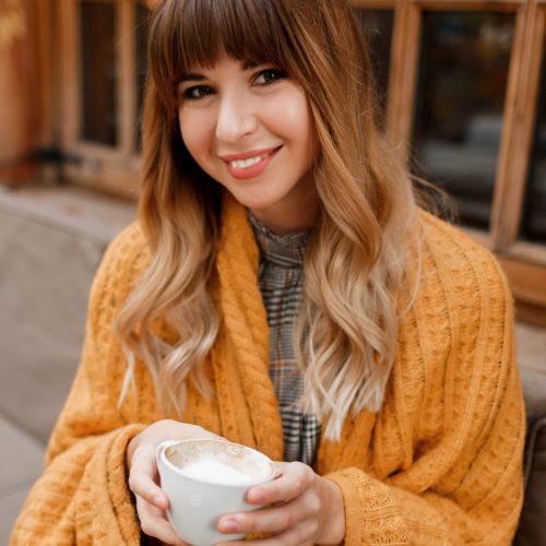 Warm cozy portrait of happy dreamy woman with wavy hairs, covered by  yellow  plaid and holding cup of hot cappuccino.  White female chilling on terrace.